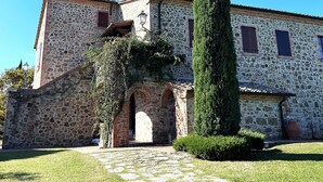 A stone building with a chimney, arched entrance, and a tall cypress tree in front. - Apartment 'Villa Tiberini 5' with Panoramic View, Shared Pool, and Wi-Fi (Torrita di Siena)
