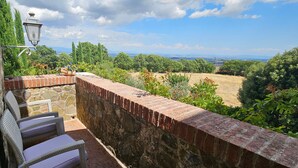 A stone wall with a brick top, wicker chairs, a potted plant, and a view of a green landscape. - Apartment 'Villa Tiberini 5' with Panoramic View, Shared Pool, and Wi-Fi (Torrita di Siena)