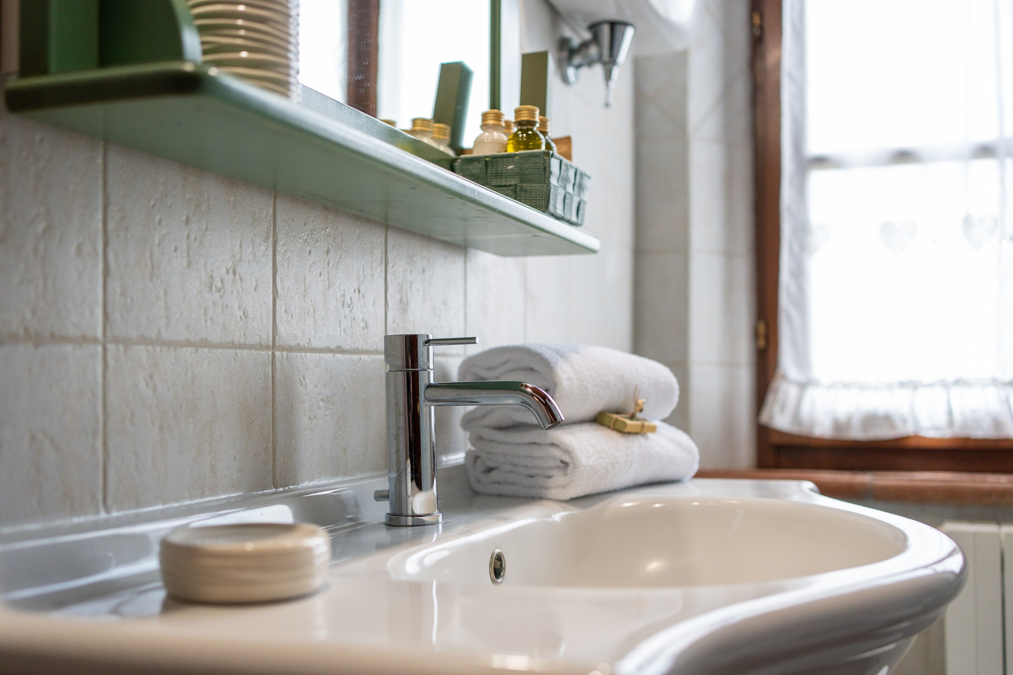 A bathroom with a white sink, a green shelf with toiletries, and a window with curtains.