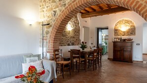 A cozy living room with a stone wall, a dining area with wooden chairs, and a kitchen with a stone archway. - Apartment 'Villa Tiberini 1' with Panoramic View, Shared Pool and Wi-Fi (Torrita di Siena)