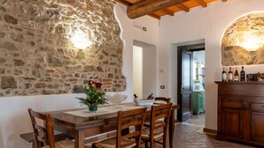 A stone wall, wooden dining table, chairs, and a kitchen area with cabinets and bottles. - Apartment 'Villa Tiberini 1' with Panoramic View, Shared Pool and Wi-Fi (Torrita di Siena)
