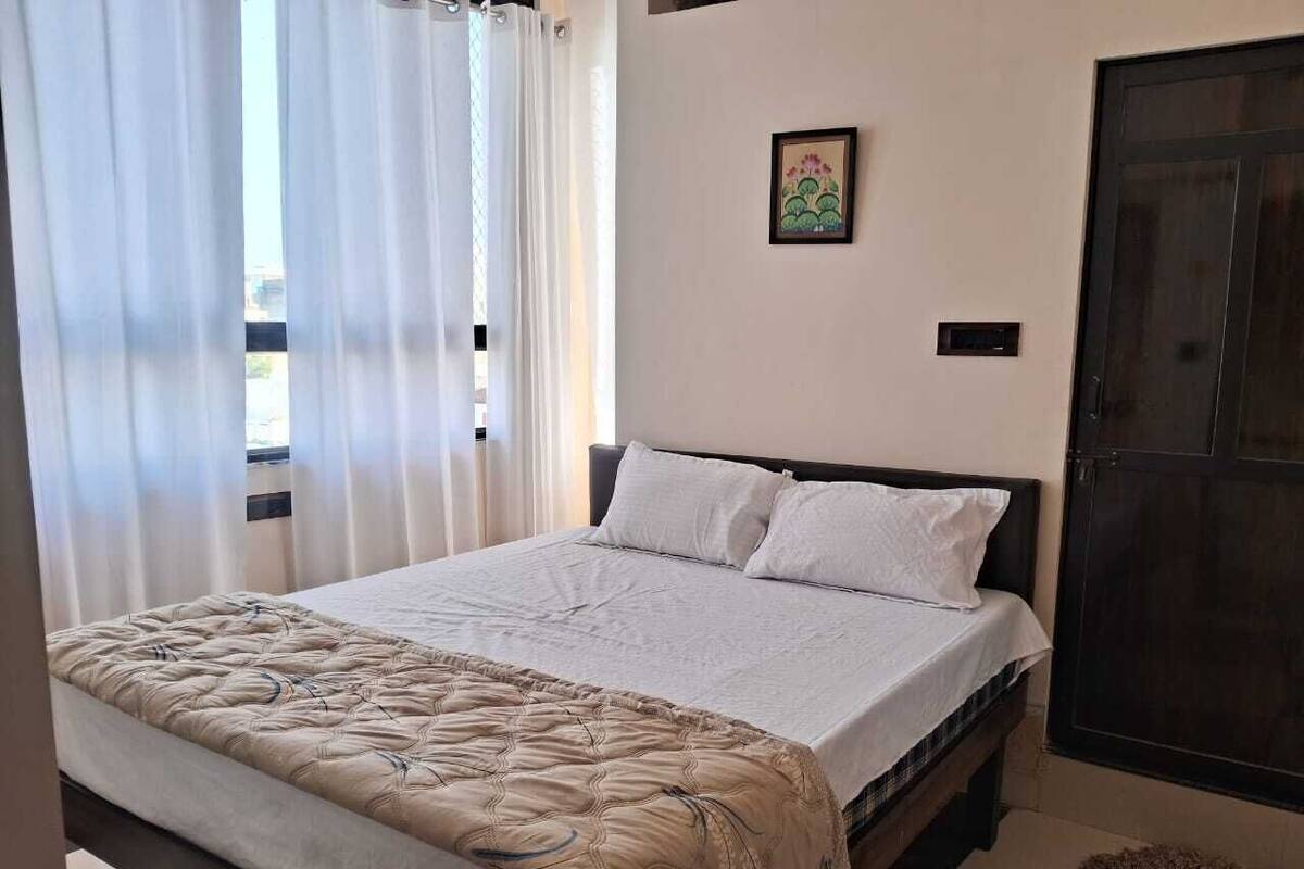 A bedroom with a bed, white curtains, a framed picture on the wall, and a dark door.