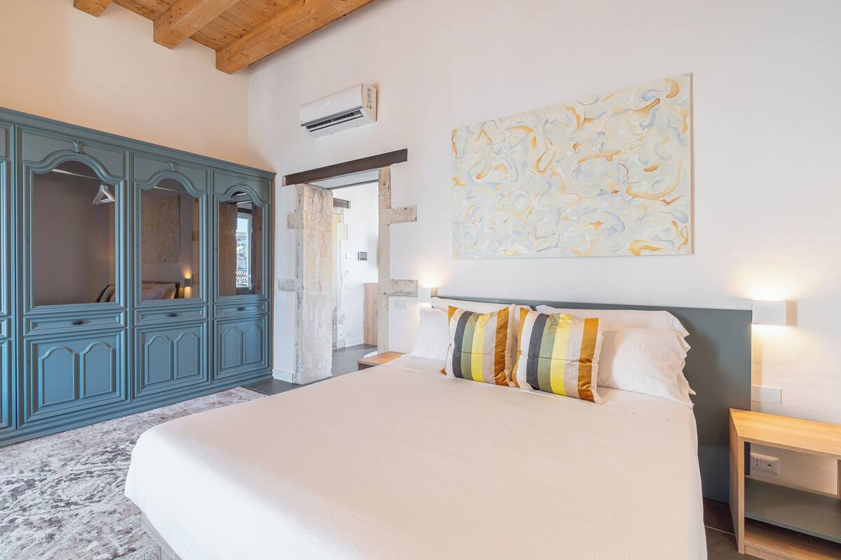 A bedroom with a large bed, a blue wardrobe, a painting on the wall, and an air conditioning unit.