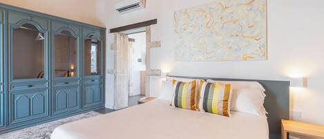 A bedroom with a large bed, a blue wardrobe, a painting on the wall, and an air conditioning unit.