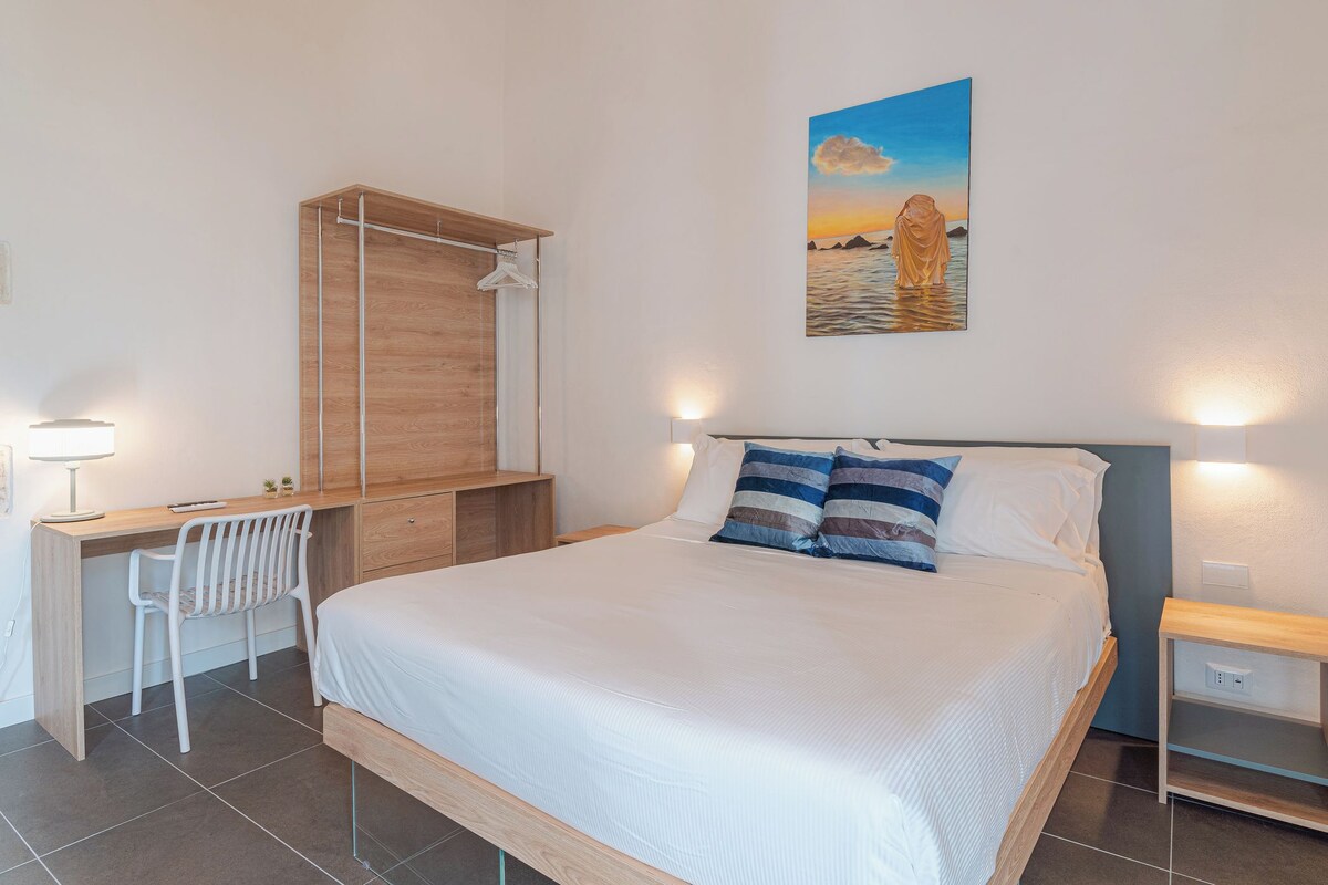 Een slaapkamer met een bed, een bureau, een stoel, een kledingkast en een schilderij aan de muur.