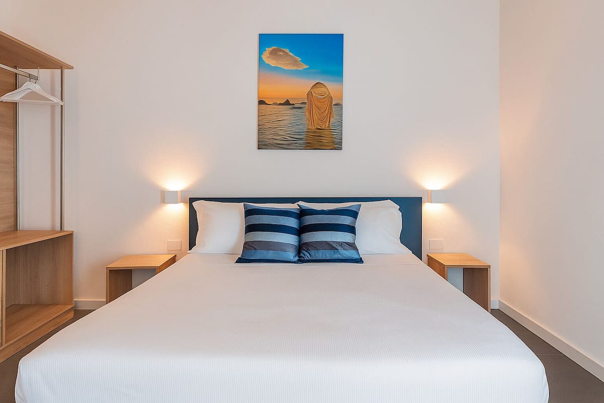 A neatly made bed with a striped pillow, two bedside tables with lamps, a wooden shelf unit, and a painting of a sunset over water on the wall.