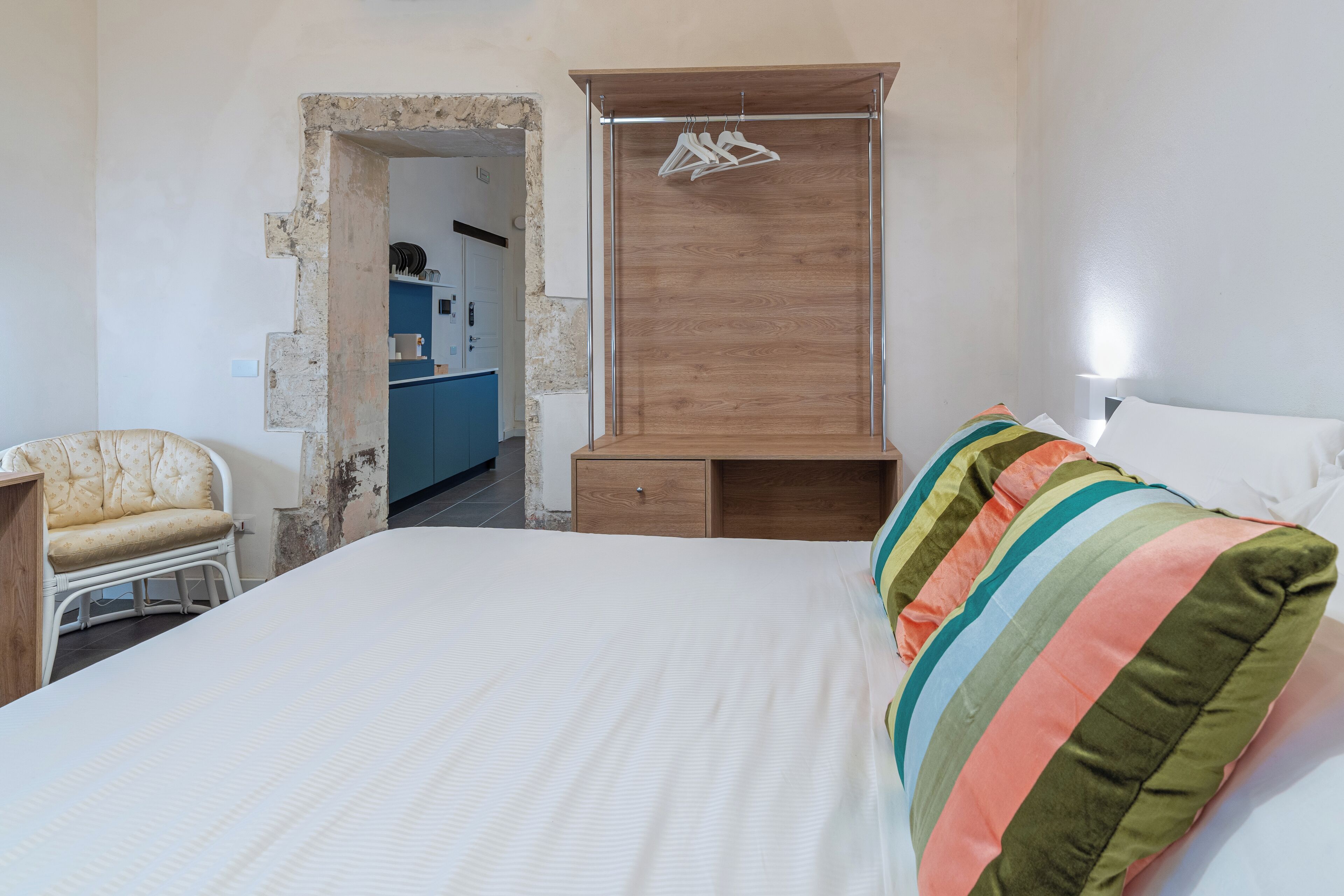 Un dormitorio con cama, armario de madera, una silla y una kitchenette al fondo.