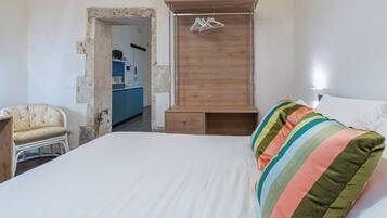 Un dormitorio con cama, armario de madera, una silla y una kitchenette al fondo.