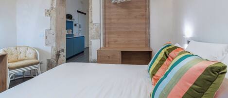 A bedroom with a bed, a wooden wardrobe, a chair, and a kitchenette in the background.