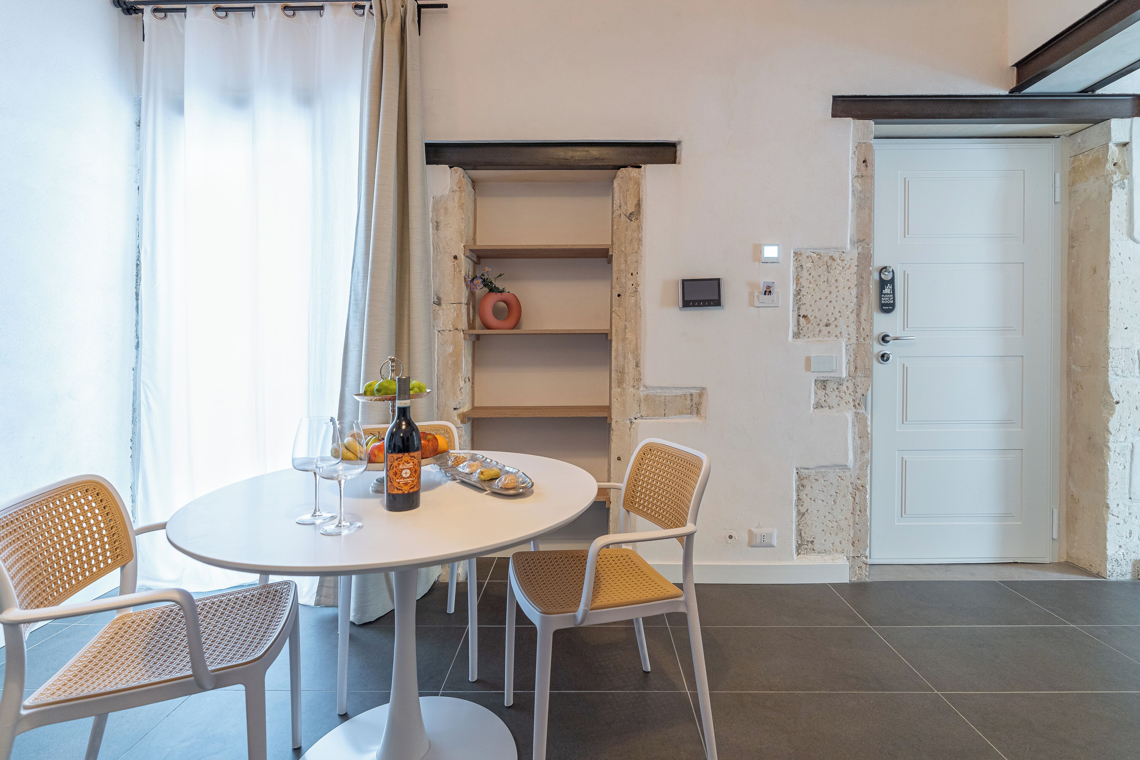 A dining area with a round table, two wicker chairs, a bottle of wine, and a fruit basket.