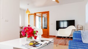 Een moderne woonkamer met een witte bank, een televisie en een eethoek met een witte tafel en een vaas bloemen.