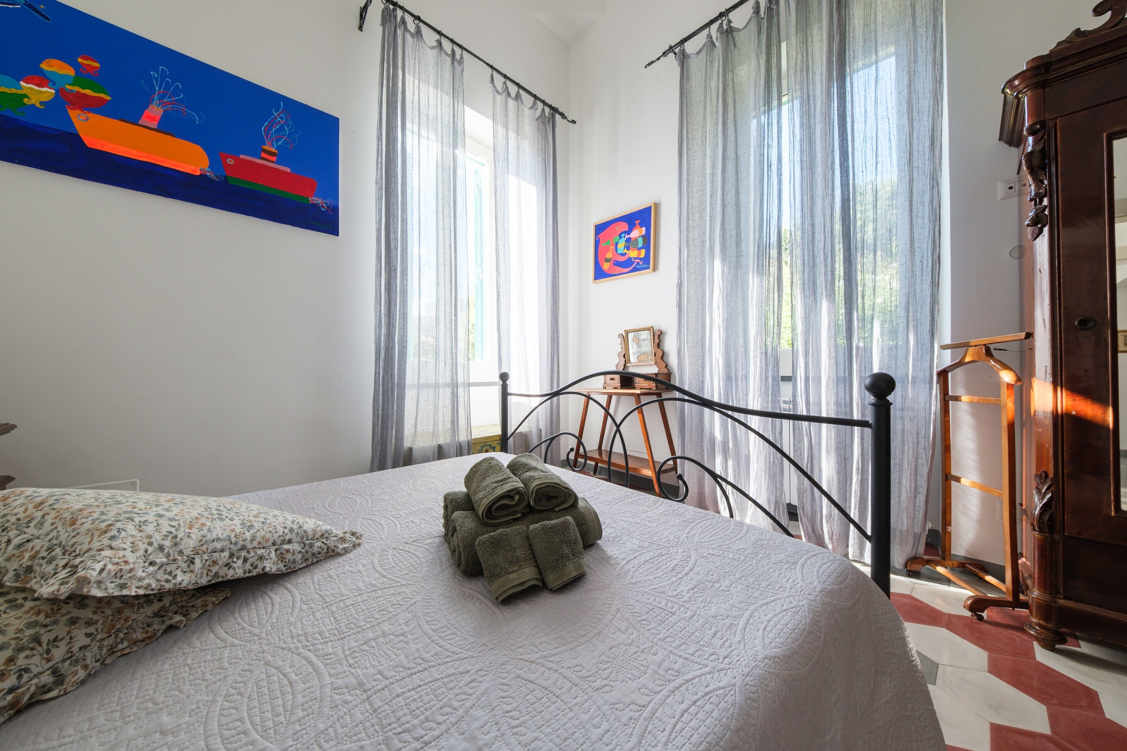 Een net opgemaakt bed met een opgerolde handdoek, een bloemenkussen en een schilderij erboven.