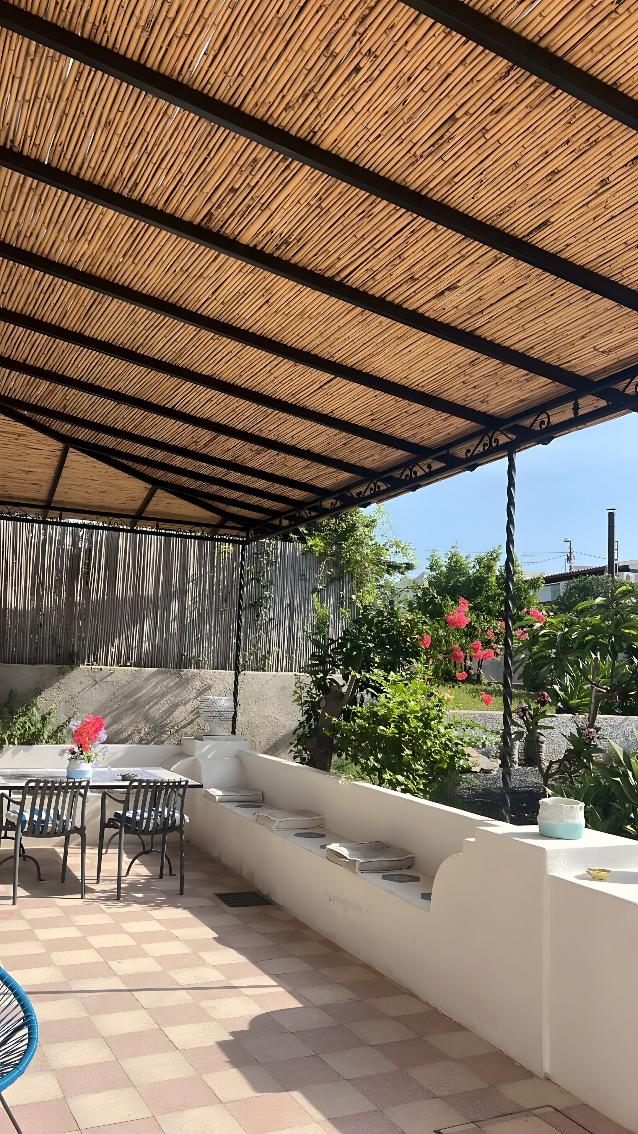 Eine Terrasse im Freien mit Bambusüberdachung, einem Tisch mit Stühlen, einer gepolsterten Bank und Topfpflanzen.