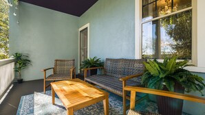 A covered outdoor seating area with a wooden table and two wicker chairs, a woven sofa, and potted plants.