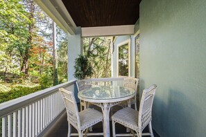 A covered outdoor area with a round glass table and wicker chairs, surrounded by a white railing and a view of trees.