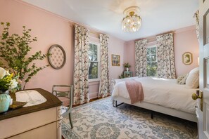 A bedroom with a bed, floral curtains, a chandelier, and a vase with flowers.