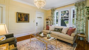 A living room with a beige sofa, a coffee table, a chandelier, and a large window with curtains. - Ever After: Luxury, Romantic Unit + Walk Downtown (Staunton)