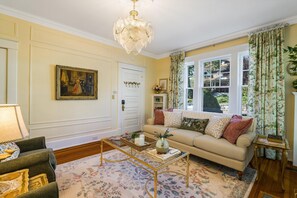 A living room with a beige sofa, a coffee table, a chandelier, and a large window with curtains.