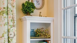 A bookshelf with decorative items and books, a floral curtain, and a window with blinds. - Ever After: Luxury, Romantic Unit + Walk Downtown (Staunton)