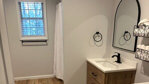 Ein Badezimmer mit einem Holzkosmetikschrank, einem Waschbecken, einem Spiegel und einem Fenster mit Jalousien.