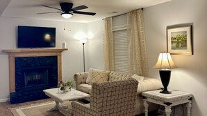 Ein Wohnzimmer mit Kamin, Deckenventilator, einer Couch, einem gemusterten Sessel, einem Couchtisch und einer Lampe.