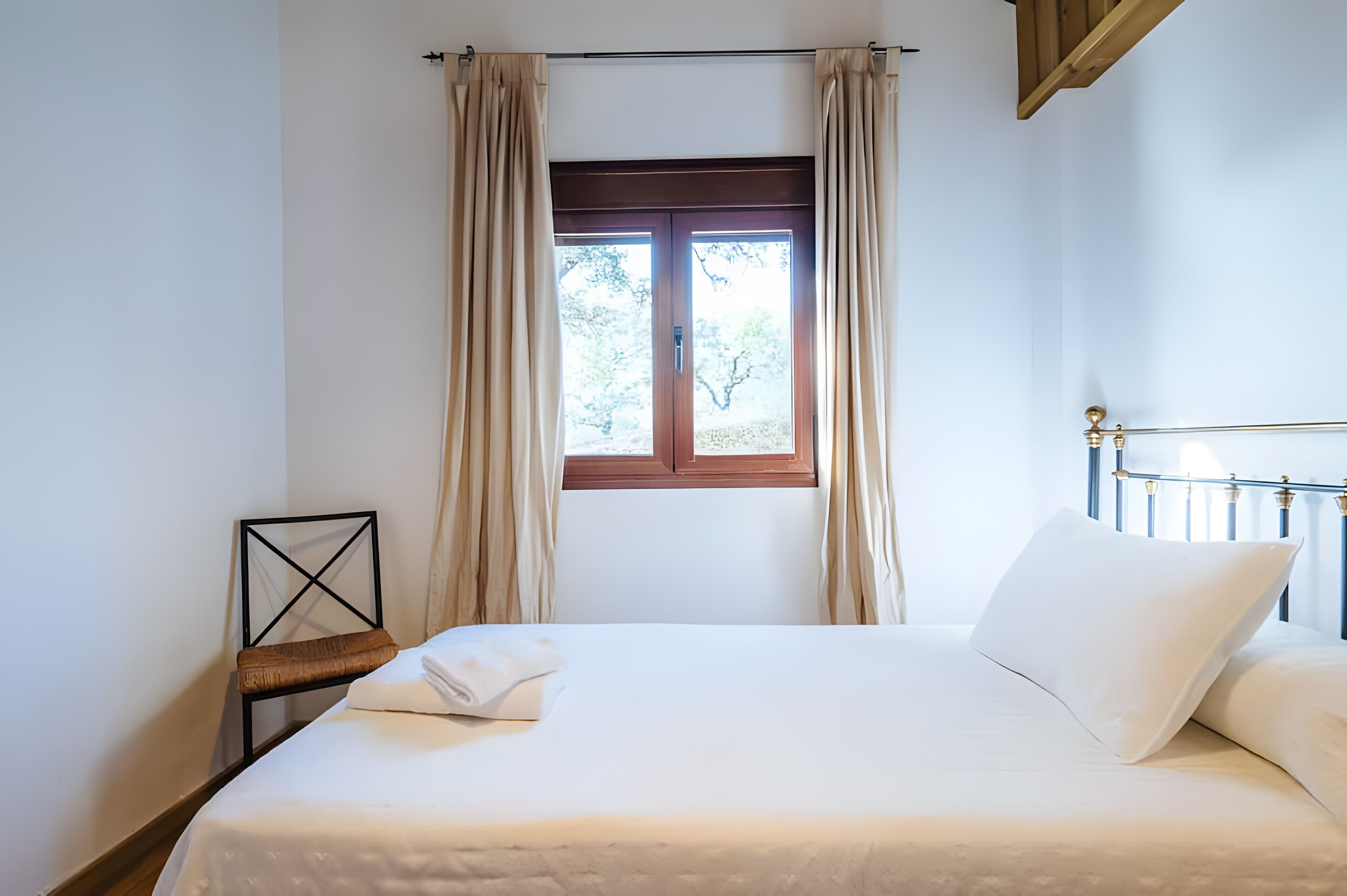 A neatly made bed with a white pillow and a folded towel, a window with curtains, and a chair with a cushion.