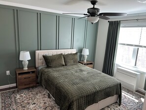 Une chambre Ă coucher avec un lit, deux tables de chevet avec des lampes, un ventilateur de plafond, une fenĂȘtre avec des rideaux et un tapis Ă motifs.