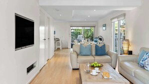Una sala moderna con un televisor de pantalla plana, un sofá color beige, una mesa de centro con manzanas y pasteles, y un comedor al fondo.
