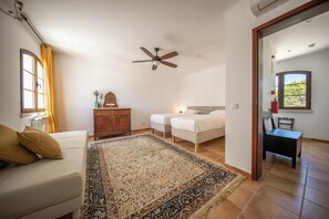 Une chambre à coucher comprenant un lit, une table de chevet, une commode, un ventilateur de plafond et une fenêtre avec des rideaux.