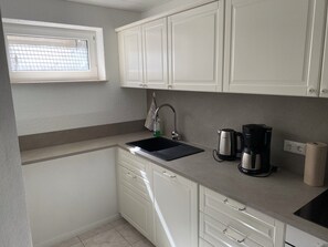 Eine kleine Küche mit weißen Schränken, einem schwarzen Spülbecken, einem Fenster, einer Kaffeemaschine und einem Wasserkocher.