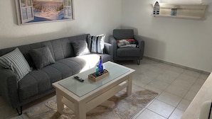 Un salon avec un canapé gris, une table basse en verre, un fauteuil gris, un tableau représentant une scène de plage et une fenêtre surmontée d’une étagère.