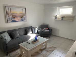 Uma sala de estar com um sofá cinzento, uma mesa de centro de vidro, uma poltrona cinzenta, um quadro com uma cena de praia e uma janela com uma prateleira acima.