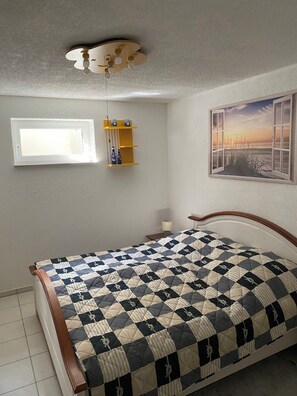 Ein Schlafzimmer mit einem Bett, einem Deckenventilator, einem Fenster mit Jalousien, einer Wandablage und einem gerahmten Bild.