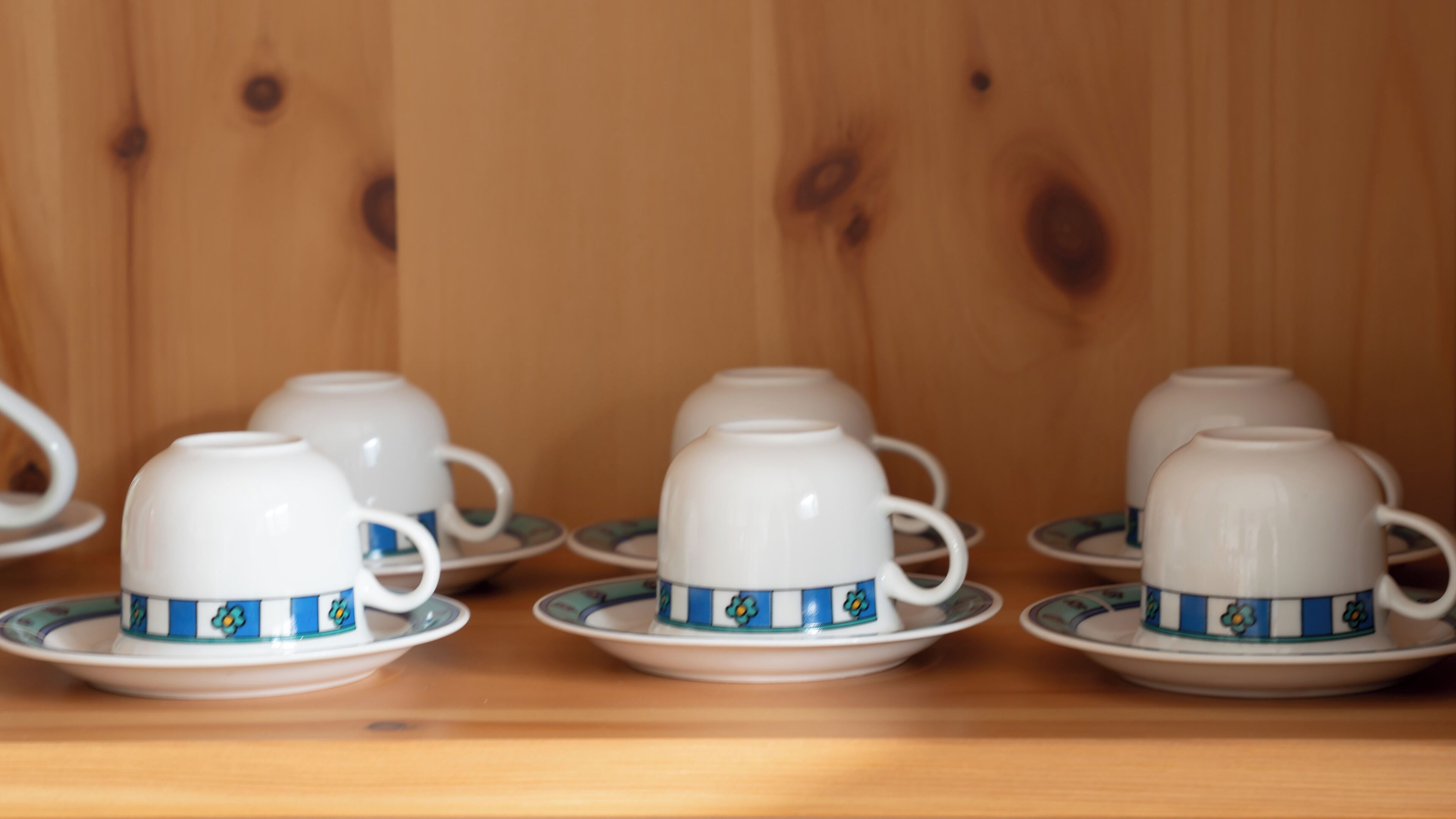 A set of white teacups with blue floral patterns on saucers, arranged on a wooden shelf.