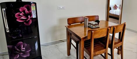 A dining area with a wooden table and chairs, a black refrigerator with floral designs, and a mirror reflecting a bed.