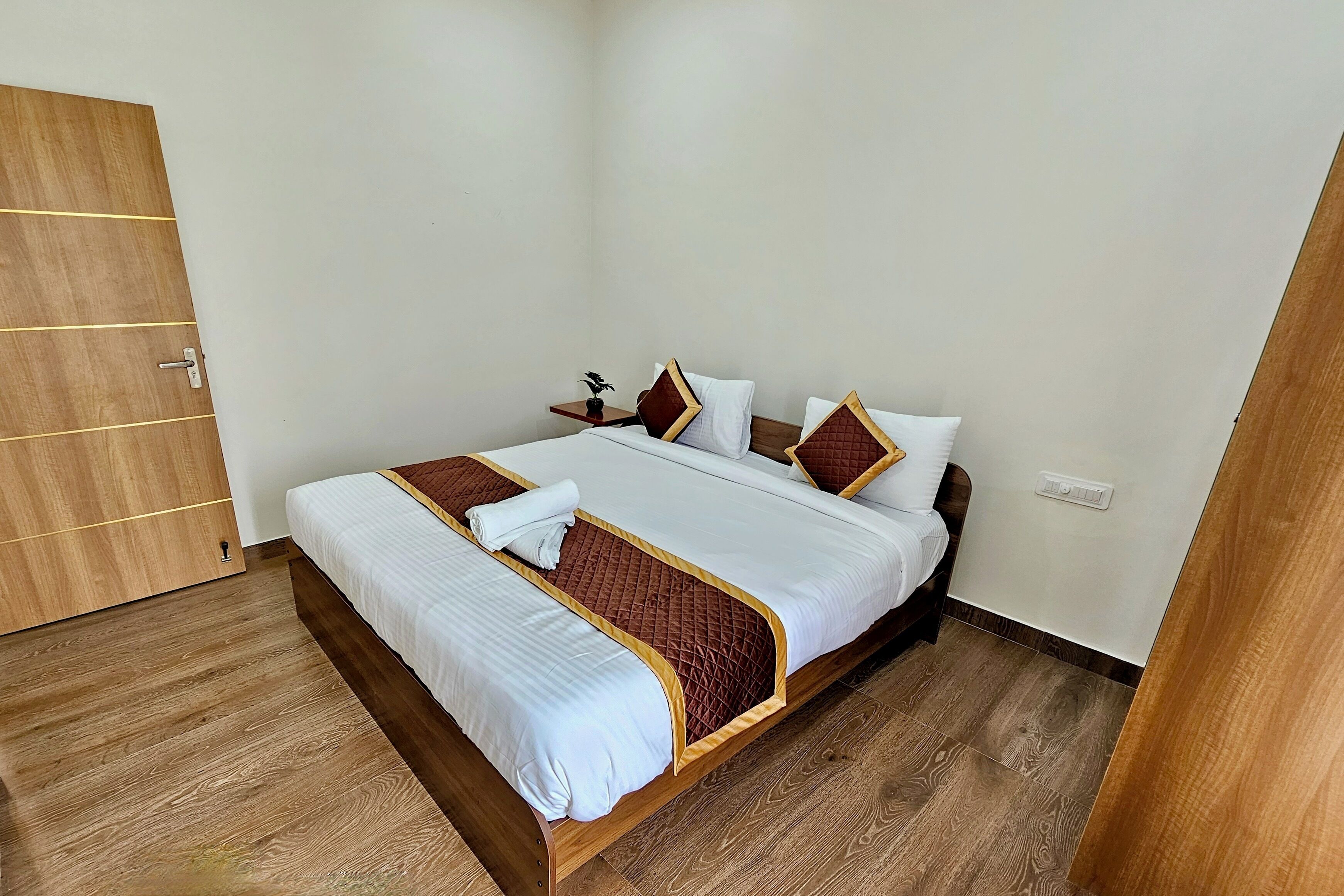 A neatly made bed with white and brown bedding, a wooden floor, and a small bedside table with a lamp.