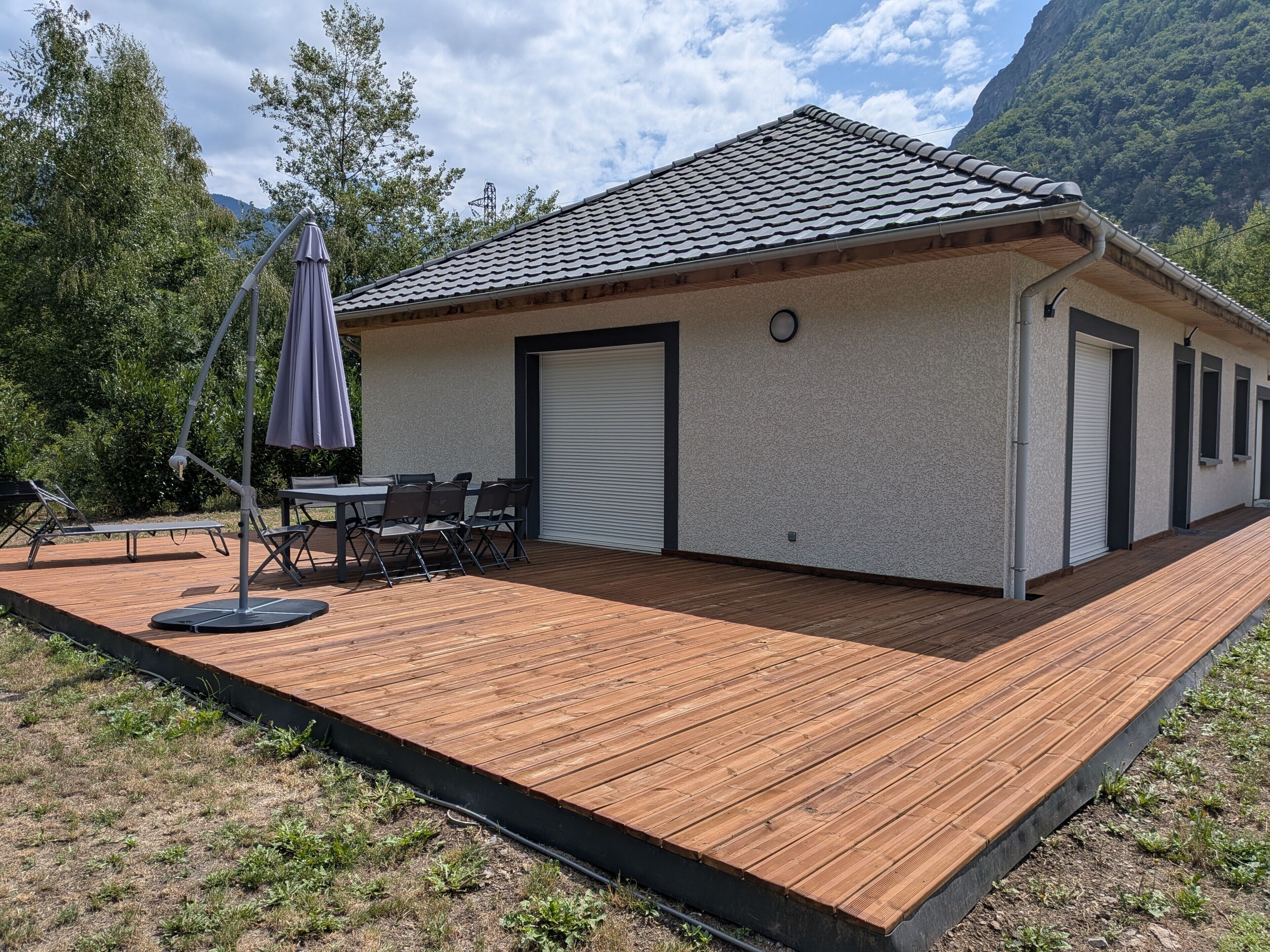 A single-story building with a tiled roof, a covered patio with a table and chairs, and a view of a mountainous landscape.