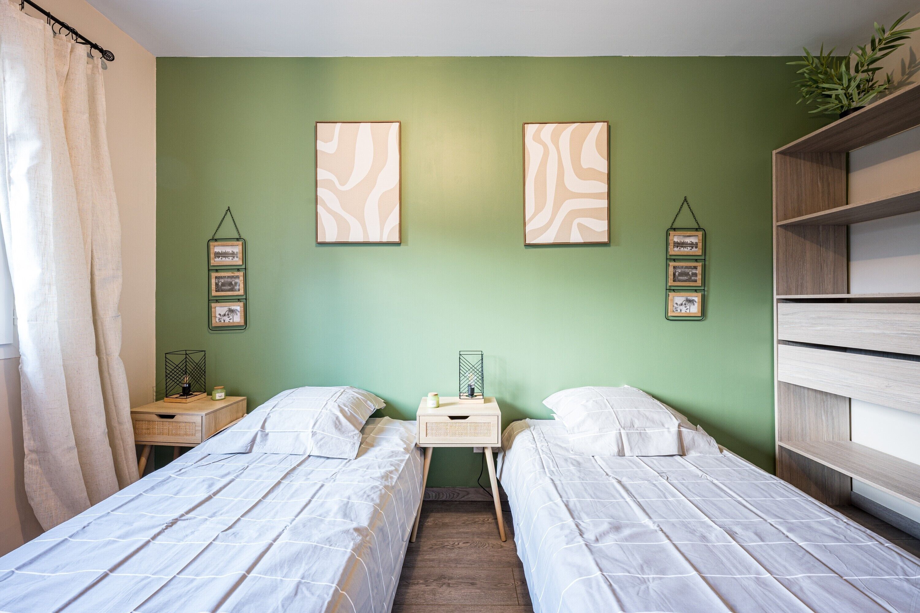 A bedroom with two single beds, a nightstand with a lamp, a small table, and a green wall with framed pictures.