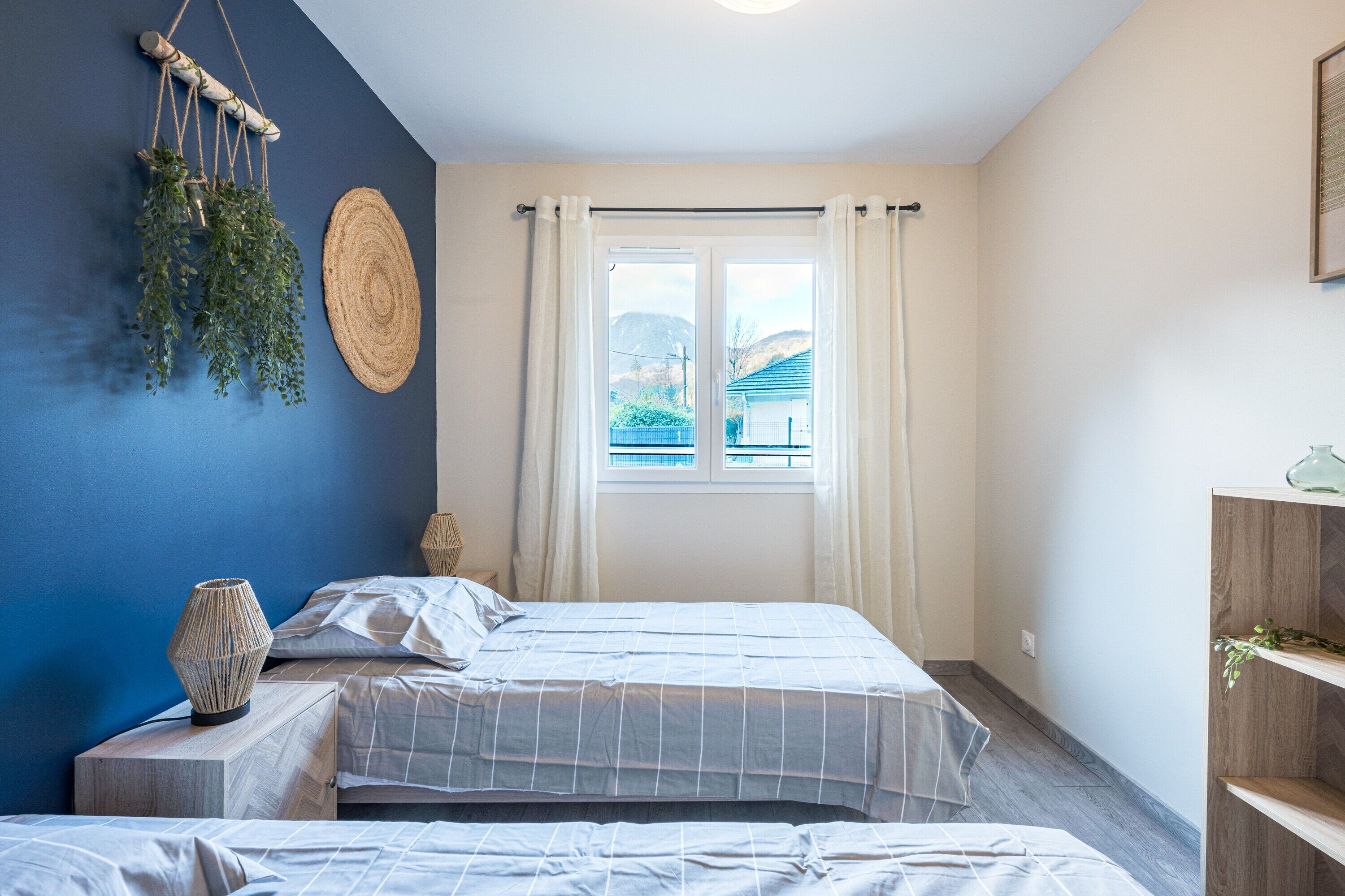 A bedroom with two beds, a window with curtains, a wall-mounted plant, and a woven lamp.