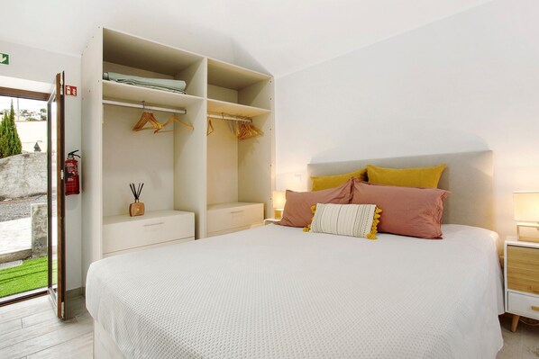 A bedroom with a bed, a wardrobe, a nightstand, and a window with a view of greenery.