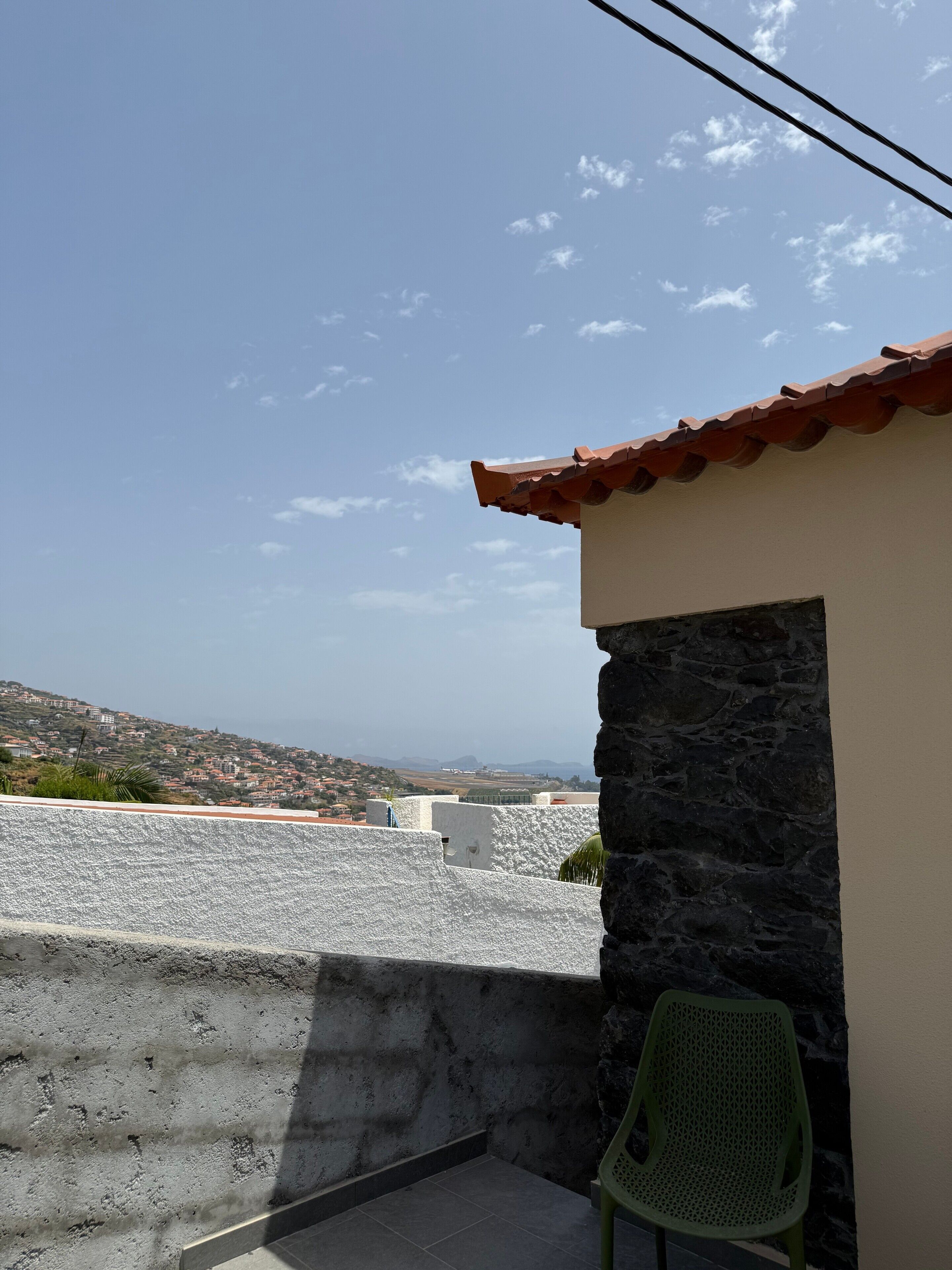 A balcony with a view of a coastal town, a stone wall, and a green chair.