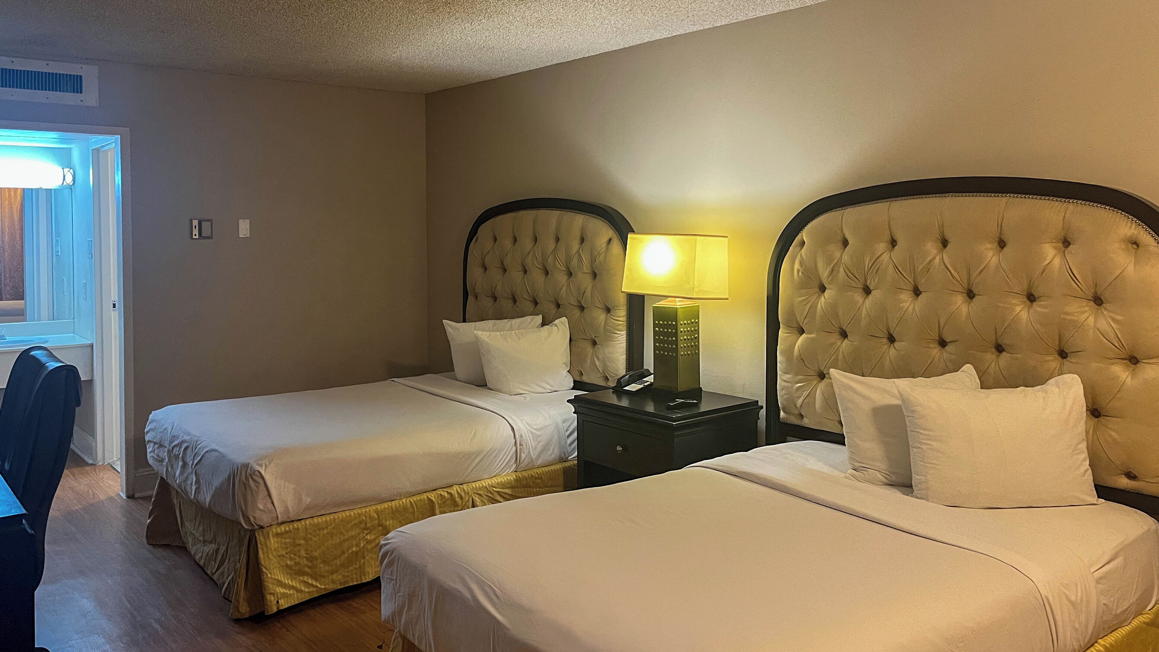 A hotel room with two beds, each with a tufted headboard, a bedside table with a lamp, and a window with blinds.