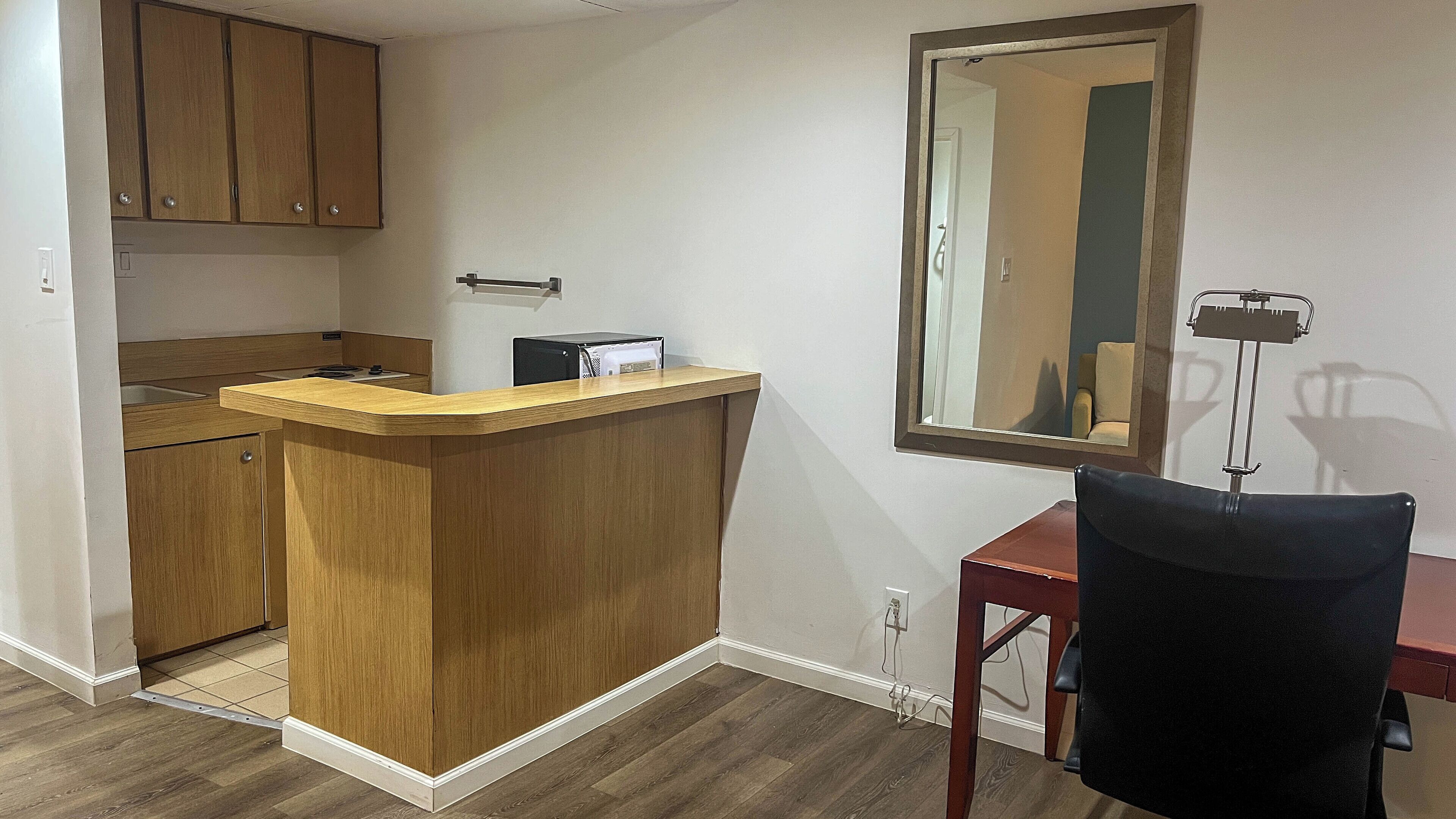 A compact kitchen with wooden cabinets, a countertop, and a mirror above a desk with a chair.
