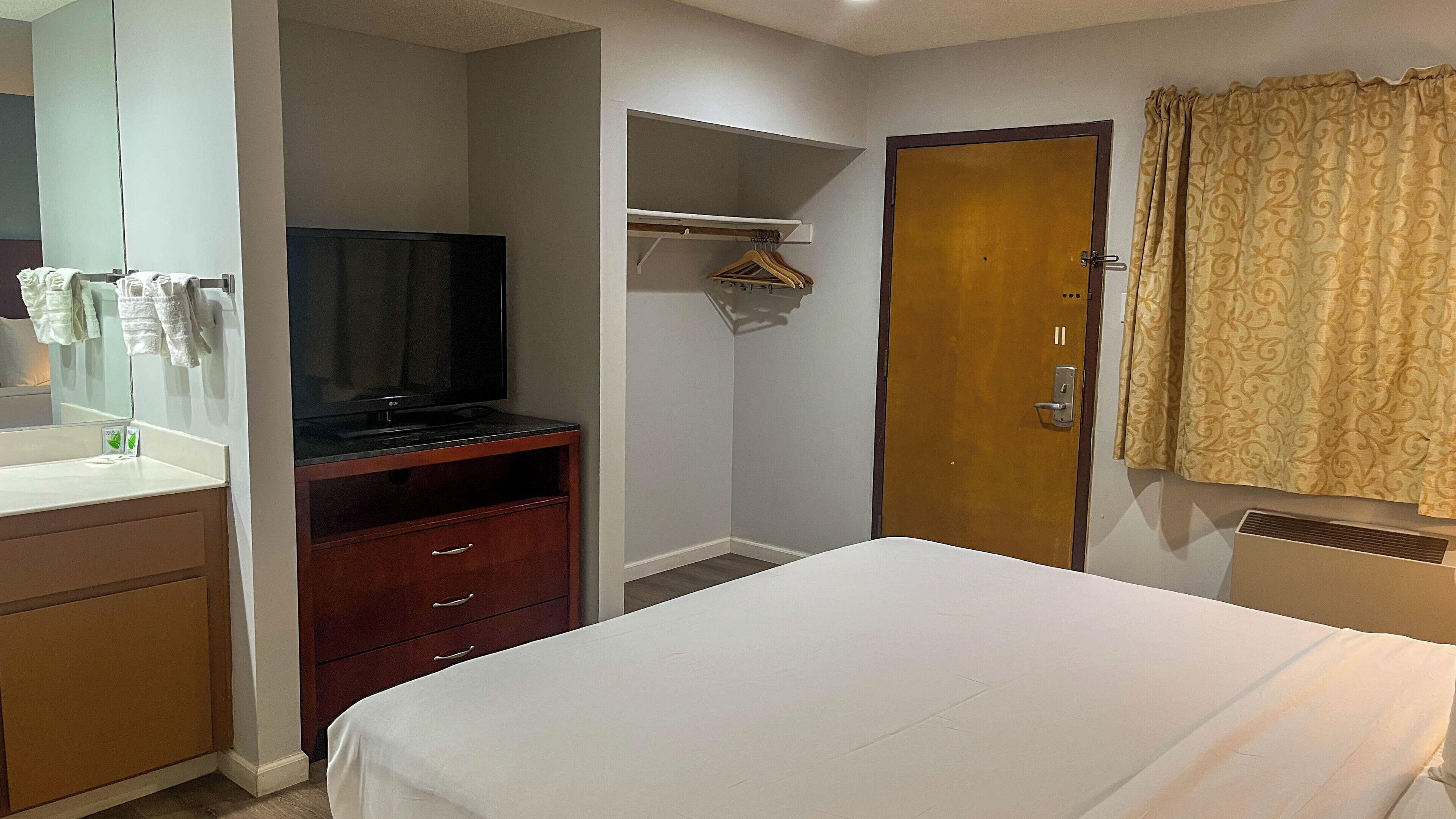 A hotel room with a bed, a TV on a wooden stand, a mirror, a wardrobe, a door, and a shower area.