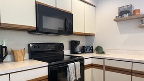 A modern kitchen with a black stove, microwave, and coffee maker. There is a wooden cutting board and a knife on the counter. - NewMont Lodge- Pool, Tennis court, pool table (Killington)
