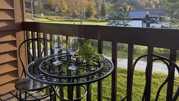 A balcony with a round metal table and chairs, overlooking a scenic view of a forest and a ski lift.