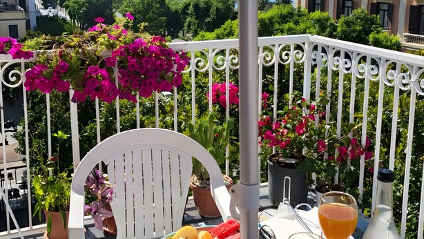Una mesa con una bandeja de fruta, dos vasos de zumo de naranja y una botella de agua en una terraza al aire libre con vistas al mar y edificios.