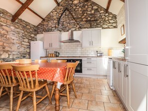 A kitchen with stone walls, white cabinets, a tiled floor, and a wooden dining table with chairs. - Ty Felin (Newport)