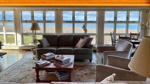 A cozy living room with a large window overlooking the beach, a sofa, a coffee table with books, and a lamp. - Amazing sunsets, a welcoming home, and island adventures.  Lifetime memories! (Chilmark)