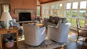 A cozy living room with a fireplace, comfortable seating, and large windows offering a view of the outdoors. - Amazing sunsets, a welcoming home, and island adventures.  Lifetime memories! (Chilmark)